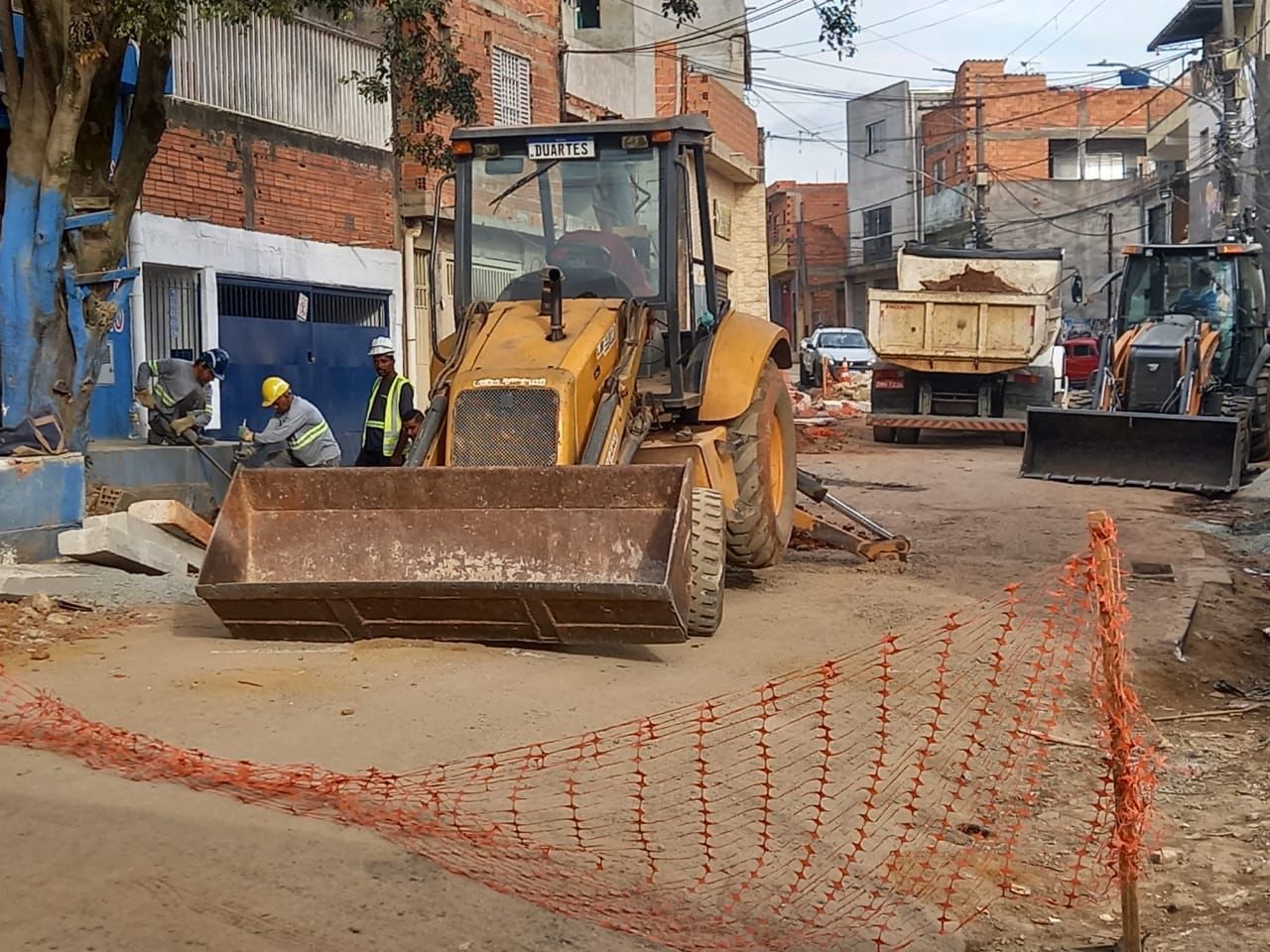 Escavadeira de cor amarela realizando a obra de construção de vias e sarjetas em uma rua de terra.